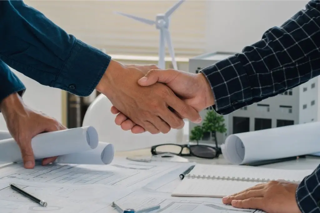 Zwei Fachleute reichen sich über technischen Bauplänen die Hand, im Hintergrund sind Modelle und Windkraftsymbole zu sehen.
